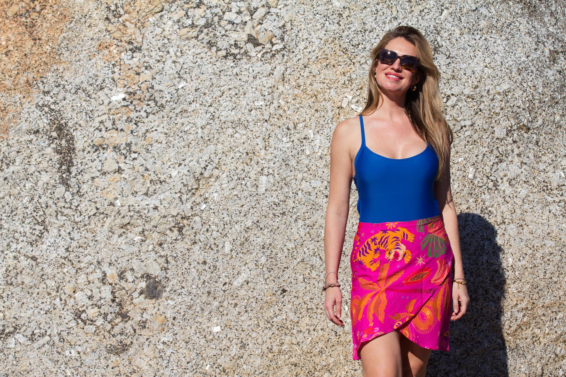 Woman wearing a blue costume and colorful pink sarong skirt standing against a textured stone rock.